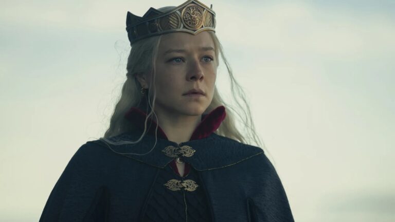 Mulher de cabelos longos e loiros usa uma coroa dourada ornamentada e um manto escuro com fechos metálicos detalhados, com parte interna vermelha visível na gola. Ela olha à frente com expressão solene contra um fundo claro de céu nublado.