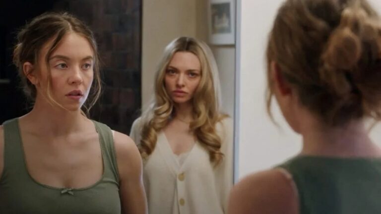 A imagem mostra duas mulheres em uma cena de tom tenso. A da esquerda, com os cabelos presos de forma desalinhada e vestindo uma blusa verde, está diante de um espelho, e sua expressão é de preocupação ou surpresa. Ao fundo, outra mulher, de cabelos loiros soltos e usando um cardigã claro, observa-a com um olhar sério. A iluminação suave e o enquadramento sugerem um momento dramático ou de confronto emocional.