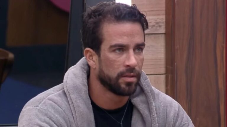 Homem com expressão séria e olhar fixo, vestindo um roupão cinza e camiseta escura por baixo, sentado em um ambiente interno com paredes de madeira e detalhes em preto. A iluminação é suave e o clima da cena parece tenso ou reflexivo.