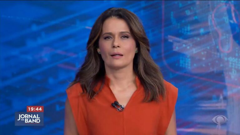 Adriana Araújo, apresentadora do Jornal da Band, aparece no estúdio do telejornal, com fundo azul e elementos gráficos. Ela veste blusa laranja e olha para a câmera com expressão séria, durante a transmissão ao vivo do noticiário das 19h44