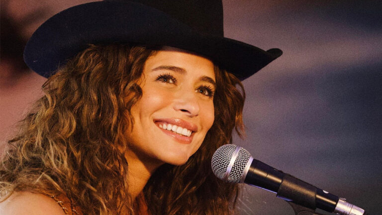 Isadora Cruz como Agrado sorri com chapéu de cowboy em frente ao microfone durante apresentação musical na novela Coração Acelerado