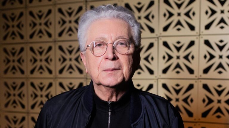 Aguinaldo Silva posa para retrato com cabelo grisalho e óculos diante de painel geométrico iluminado em ambiente interno