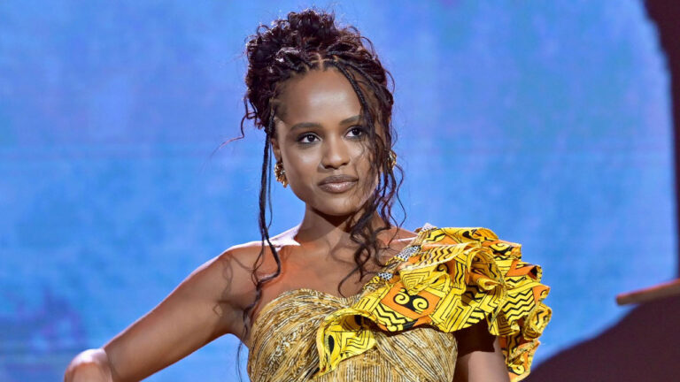 Duda Santos usa vestido amarelo estampado e cabelos presos com tranças soltas em evento da Globo com fundo azul desfocado