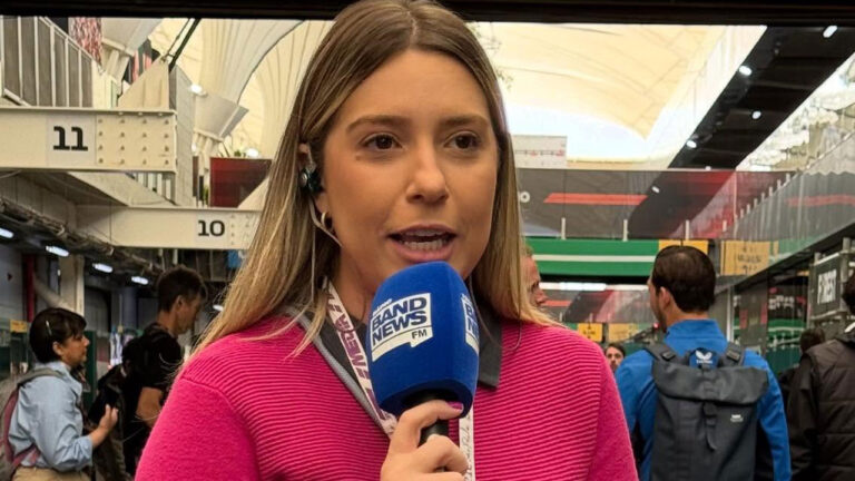 Alinne Fanelli fala ao microfone da BandNews FM em área de cobertura com boxes de autódromo e pessoas ao fundo durante reportagem