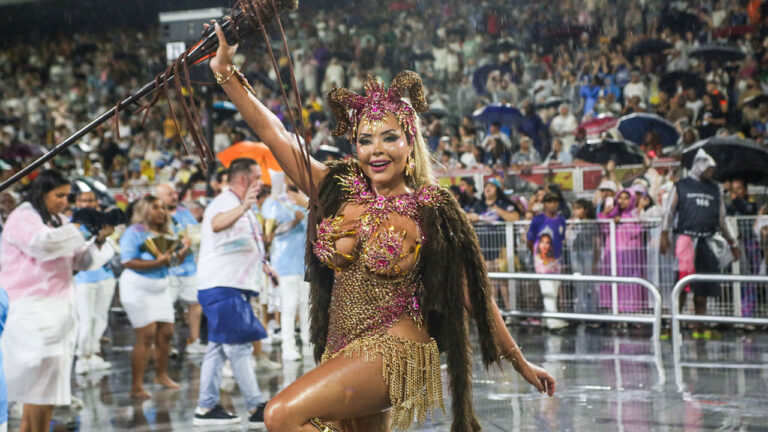 Ana Beatriz Godoi desfila com fantasia luxuosa de chifres e brilhos segurando um cetro sob chuva no Sambódromo durante o Carnaval