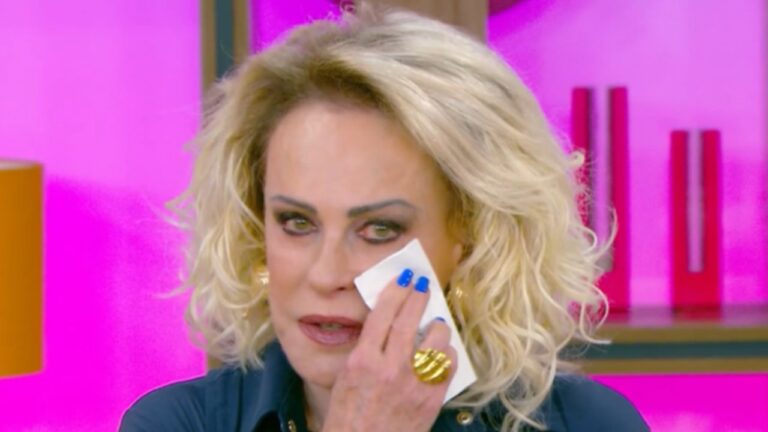 Mulher de cabelos loiros e cacheados enxuga o rosto com um lenço de papel, aparentando emoção. Ela usa esmalte azul vibrante, anel dourado grande e blusa escura. O fundo colorido em tons de rosa e laranja sugere um estúdio de televisão iluminado.