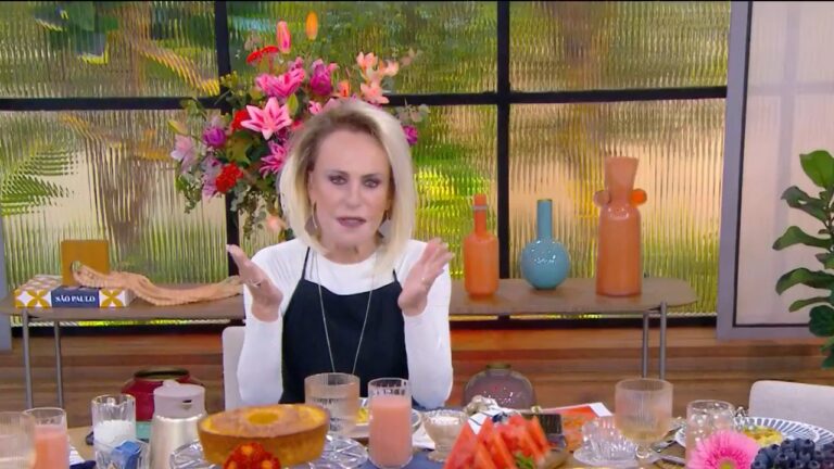 Mulher apresenta programa matinal sentada à mesa com café da manhã servido, incluindo bolo, sucos e frutas; ao fundo, há vasos coloridos e arranjo de flores sobre aparador próximo à janela.