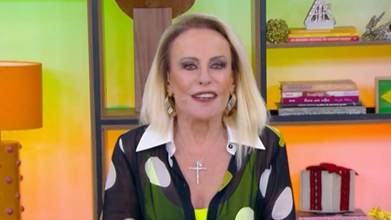 Mulher de cabelos loiros e lisos usa blusa preta transparente com bolinhas brancas e verdes, colar prateado em formato de cruz e maquiagem marcada nos olhos, sorrindo diante de uma estante colorida com livros e objetos de decoração ao fundo, incluindo uma pequena bandeira do Brasil e um abajur laranja à esquerda