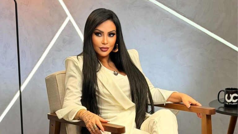 Ana Paula Oliveira sentada em poltrona de estúdio com cenário geométrico ao fundo usando conjunto branco e cabelo longo preto solto