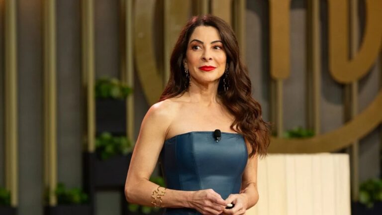 Mulher de cabelos castanhos longos e ondulados está em um estúdio bem iluminado, vestindo um vestido tomara que caia azul escuro. Ela usa batom vermelho, brincos longos e pulseira dourada, com expressão serena enquanto olha para o lado, tendo ao fundo uma parede com detalhes dourados e plantas.