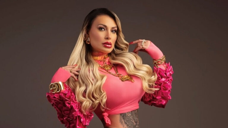 Andressa Urach posa para foto promocional com top rosa mangas volumosas joias douradas e cabelo loiro longo em estúdio neutro