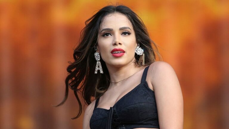 Anitta, com cabelos soltos e maquiagem marcante, está em frente a um fundo alaranjado, usando um top preto e brincos grandes decorativos — um deles em formato de letra “A”. A iluminação e o enquadramento sugerem que ela está se apresentando em um palco