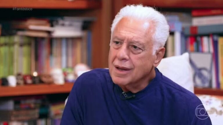 Homem de cabelos brancos e expressão séria usa blusa azul durante entrevista em ambiente com estante de livros ao fundo