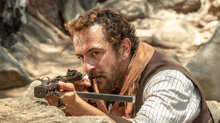 Irandhir Santos como Arduíno aponta uma arma e morde um lenço em cena da novela Guerreiros do Sol em meio a pedras no sertão