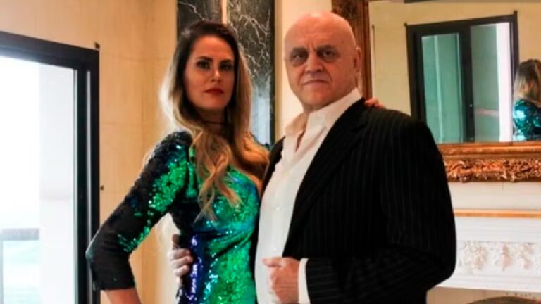 Homem e mulher posam lado a lado em um ambiente sofisticado com molduras douradas e paredes claras. A mulher usa um vestido justo com paetês em tons de verde e azul, cabelos soltos e expressão séria. O homem veste terno preto listrado com camisa branca sem gravata, mantendo uma postura confiante e olhar direto para a câmera.