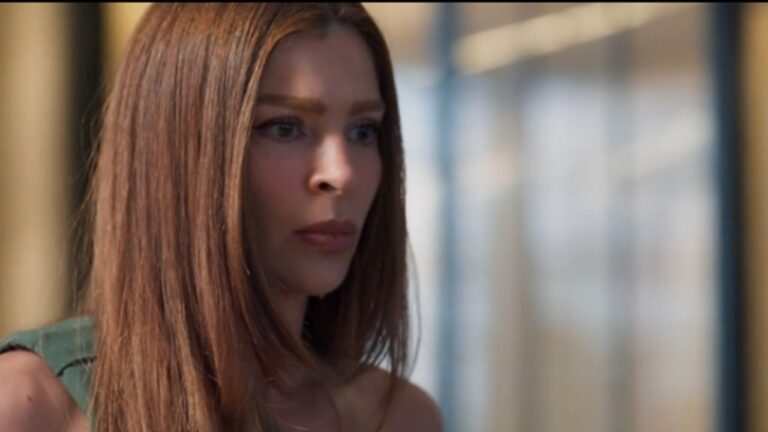 Mulher com expressão séria olha fixamente para frente em ambiente interno com iluminação suave e fundo desfocado.