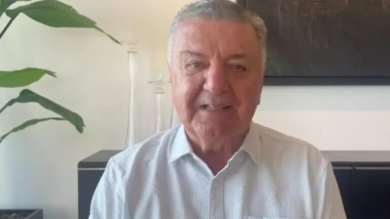 Homem idoso sorri levemente em ambiente interno iluminado. Ele veste camisa social clara e aparece diante de uma parede branca com um quadro escuro ao fundo. À esquerda, há uma planta verde em vaso e objetos de decoração sobre um móvel preto