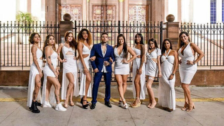 Arthur O Urso posa de terno azul cercado por suas nove mulheres vestidas de branco em frente a uma grade de ferro e um casarão