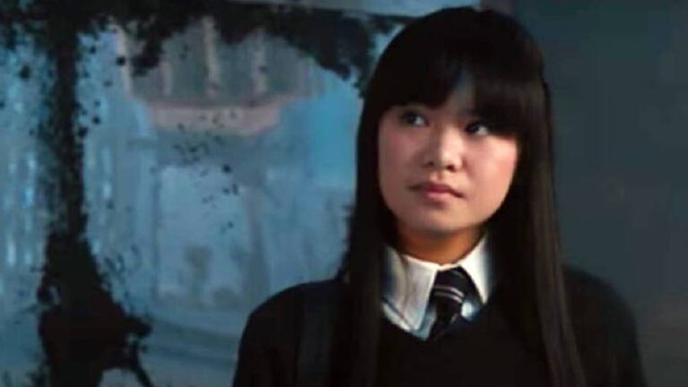 Jovem com expressão séria usa uniforme escolar preto e branco com gravata, em ambiente interno com fundo azul e iluminação suave.