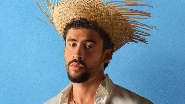 Homem com barba e cabelo cacheado usa chapéu de palha e camisa clara em retrato com fundo azul olhando para a câmera