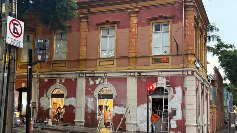 A imagem mostra um prédio antigo de esquina, com arquitetura clássica e fachada em tons de vermelho e amarelo. A pintura está sendo restaurada, com partes das paredes remendadas e andaimes apoiados. Há pessoas trabalhando próximas às portas, e um sinal de pedestre vermelho aceso no canto da rua. Um letreiro digital exibe “ALOS” e há uma placa de trânsito indicando proibição de estacionamento.