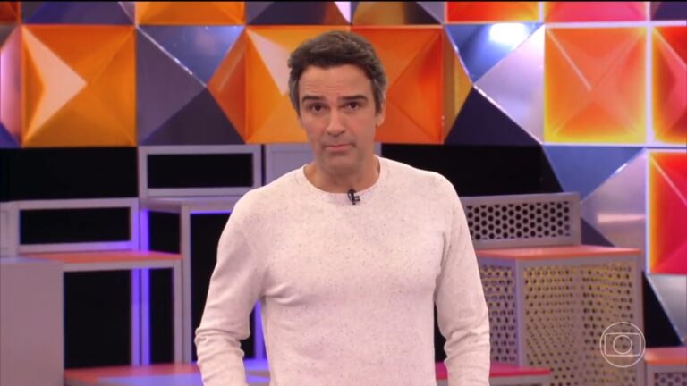 Tadeu Schmidt, apresentador do BBB 26 aparece em estúdio colorido da TV Globo, com painéis geométricos em tons de laranja, amarelo e roxo ao fundo. Ele usa suéter claro e microfone de lapela, olhando para a câmera com expressão séria