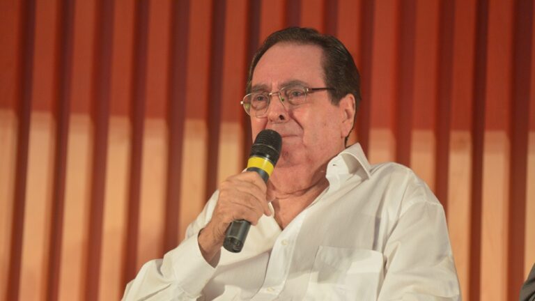 Homem segura microfone e fala em evento público vestindo camisa branca diante de painel com ripas de madeira ao fundo