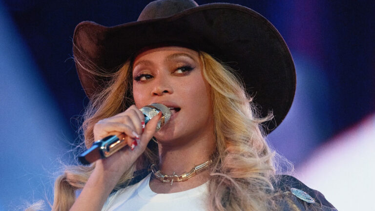 Beyoncé canta ao microfone com chapéu de cowboy marrom e cabelo loiro ondulado durante show da era Cowboy Carter sob luzes azuis