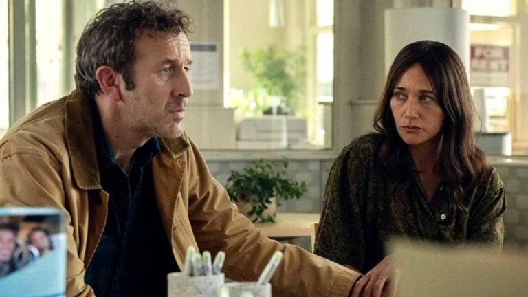 Um homem e uma mulher estão sentados lado a lado em um ambiente que parece um escritório ou sala de espera. Ambos têm expressões sérias e parecem estar tendo uma conversa tensa ou preocupada. O homem usa uma jaqueta marrom clara e olha para frente, enquanto a mulher, de cabelos escuros soltos e blusa estampada, o observa atentamente. O cenário é iluminado por luz natural que entra pelas janelas ao fundo.