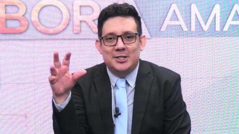 Erlan Bastos, vestindo terno preto, camisa azul e gravata clara fala de forma expressiva em estúdio de TV, gesticulando com a mão levantada. Ao fundo, um painel eletrônico exibe parcialmente o nome do jornal Bora Amapá