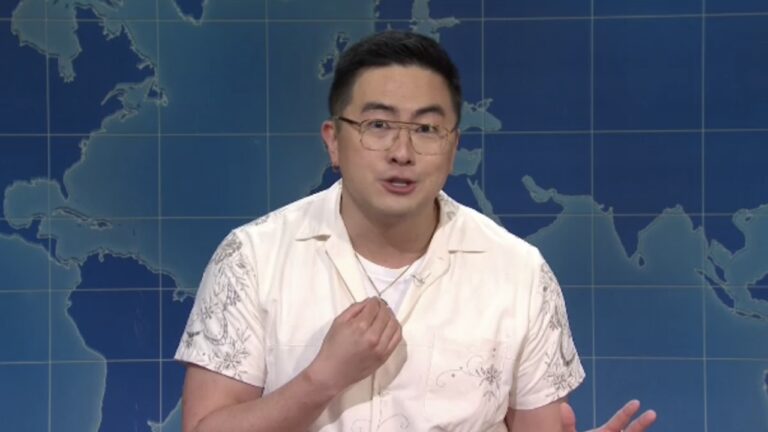 Homem fala em frente a um fundo azul com mapa-múndi em linhas brancas, vestindo camisa clara estampada e óculos de armação dourada, com uma das mãos levantada e a outra próxima ao peito enquanto faz expressão de explicação.