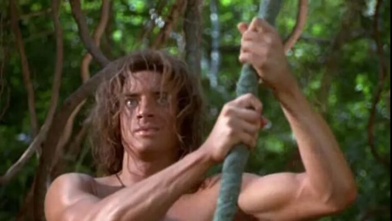 Homem de aparência selvagem, com cabelos longos e desgrenhados, segura uma grossa videira com as duas mãos em meio a uma floresta tropical. Ele está sem camisa, exibindo músculos definidos, e observa algo à frente com expressão determinada enquanto se apoia na corda cercado por vegetação verde e densa.