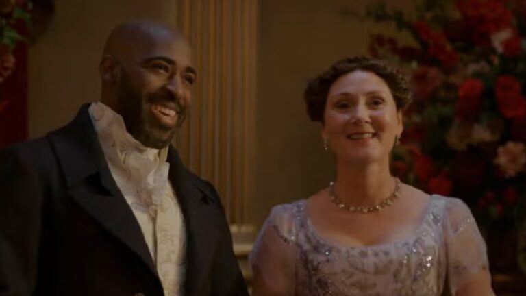 Homem e mulher com trajes de época sorriem lado a lado em salão elegante decorado com flores e iluminação quente