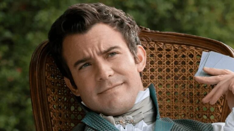 Homem de expressão pensativa reclina-se em cadeira de palha ao ar livre, vestindo roupa de época com colete azul e lenço branco no pescoço, segurando um livro na mão.