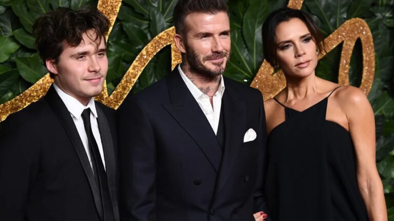 Três pessoas posam lado a lado em evento de gala usando trajes formais em frente a painel verde com folhas e elementos dourados homem jovem à esquerda usa terno preto camisa branca e gravata preta tem cabelos castanhos ondulados e expressão discreta homem ao centro usa terno preto de abotoamento duplo camisa branca sem gravata lenço branco no bolso barba bem aparada cabelos curtos e penteados para cima com expressão confiante mulher à direita usa vestido preto de alça fina cabelos castanhos presos maquiagem marcada e postura elegante todos olham levemente para fora da câmera em ambiente iluminado típico de tapete vermelho