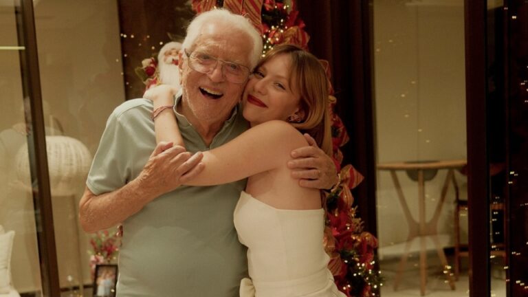 Homem idoso e mulher jovem se abraçam sorrindo em frente a árvore de Natal decorada