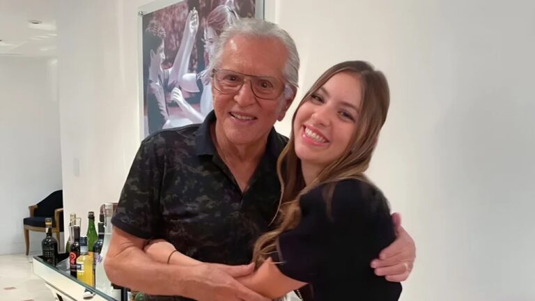 Homem idoso e mulher jovem sorriem enquanto se abraçam em ambiente interno bem iluminado. O homem usa camisa preta estampada e óculos de armação quadrada, e a mulher veste blusa preta com cabelo longo e solto. Ao fundo há uma prateleira com garrafas de bebidas, uma cadeira e uma grande fotografia emoldurada na parede mostrando pessoas em um momento descontraído.