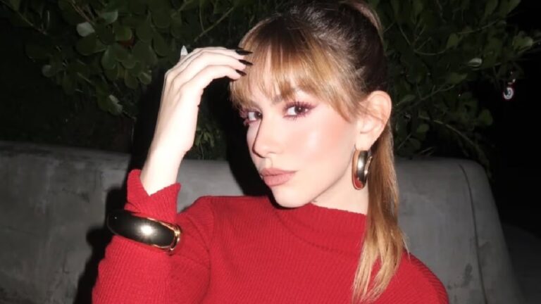 Mulher posa à noite ao ar livre usando blusa vermelha de gola alta, brincos grandes dourados e pulseira metálica. Ela tem franja e cabelo preso em rabo de cavalo, maquiagem destacando os olhos e os lábios, e segura uma mecha de cabelo com uma das mãos enquanto olha para a câmera.