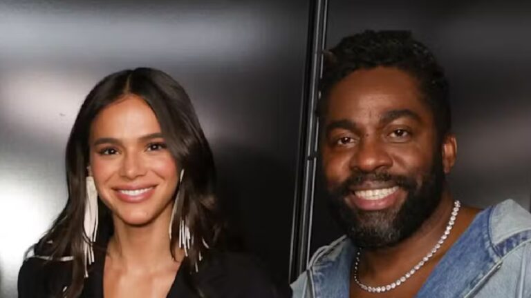 Homem e mulher posam sorridentes lado a lado em ambiente interno com fundo escuro; ela usa brincos longos e roupa preta, e ele veste jaqueta jeans e colar prateado.