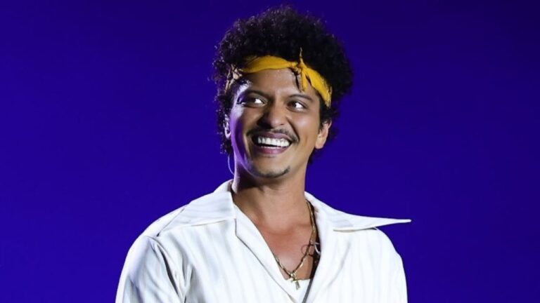 Homem sorri em frente a um fundo roxo iluminado, usando camisa branca com gola aberta, colares dourados e faixa amarela amarrada na cabeça. Ele parece estar em um palco, sob luzes de show.
