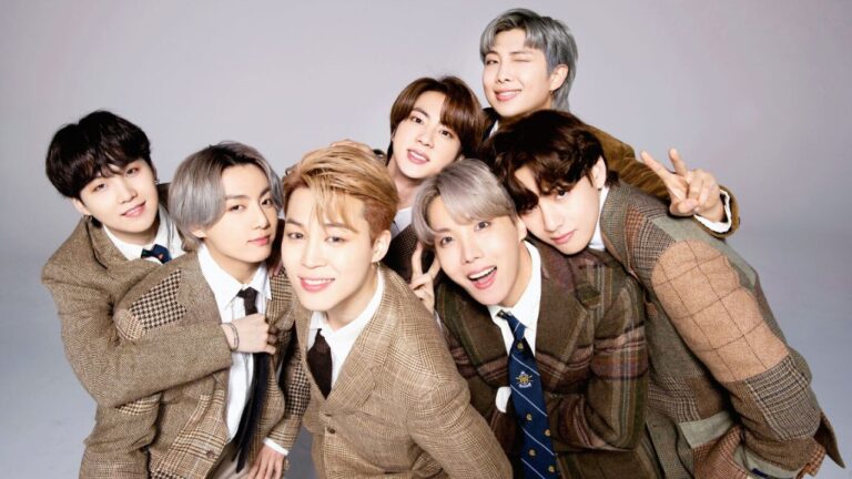 Sete jovens posam sorrindo juntos para a foto, todos usando ternos marrons em diferentes tons e padrões de xadrez. Eles estão próximos uns dos outros, com expressões alegres, e um deles faz o gesto de paz com os dedos. O fundo é neutro e claro, destacando o grupo.