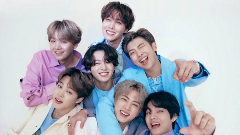 Sete jovens posam juntos sorrindo em fundo claro, vestindo roupas coloridas em tons de azul, lilás e creme, abraçados e com expressões alegres e descontraídas voltadas para a câmera.