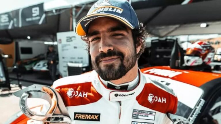 Piloto de corrida com macacão branco e vermelho e boné azul segura troféu sorrindo em área de boxes