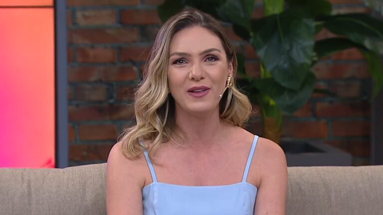 Foto de Camila Galetti sentada em sofá de estúdio televisivo sorri enquanto fala, usando um vestido azul-claro de alças finas e microfone de lapela na orelha. Ao fundo, há uma parede de tijolos aparentes e plantas decorativas, compondo o cenário do programa Mulheres, da TV Gazeta