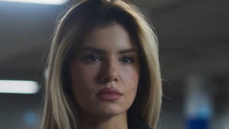 Mulher loira em close observa à frente com expressão séria em ambiente interno com iluminação fria e fundo desfocado