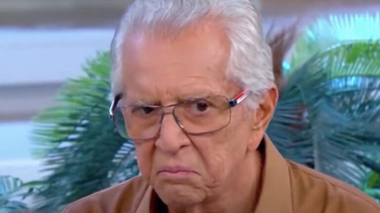 Carlos Alberto de Nóbrega faz expressão de descontentamento usando óculos de grau e camisa marrom com folhagens verdes ao fundo