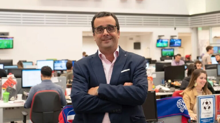 Carlos Maluf de braços cruzados sorri na redação da ESPN em São Paulo com diversos profissionais e monitores de TV ao fundo da imagem
