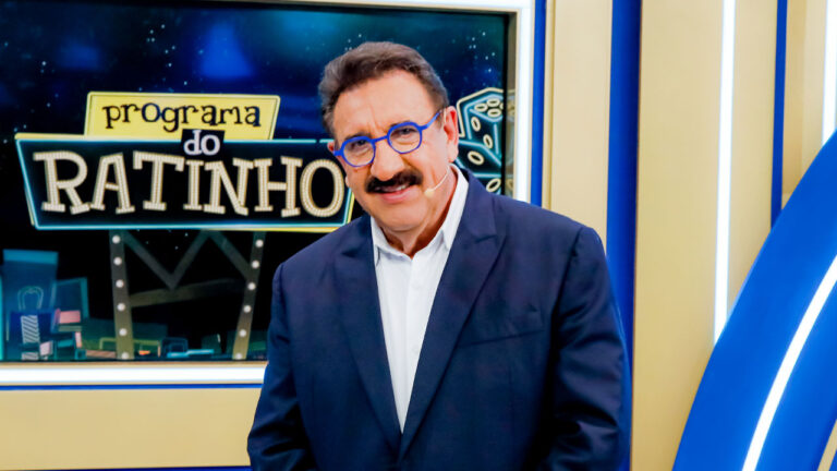 Carlos Massa o Ratinho de terno azul e óculos de armação colorida sorri no cenário de seu programa com o logotipo da atração ao fundo no SBT
