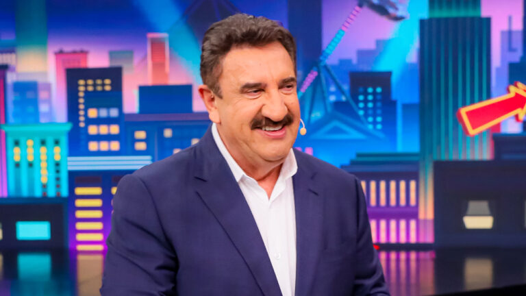 Carlos Massa, o Ratinho, sorri usando terno azul e camisa branca no cenário do Programa do Ratinho exibido na programação do SBT