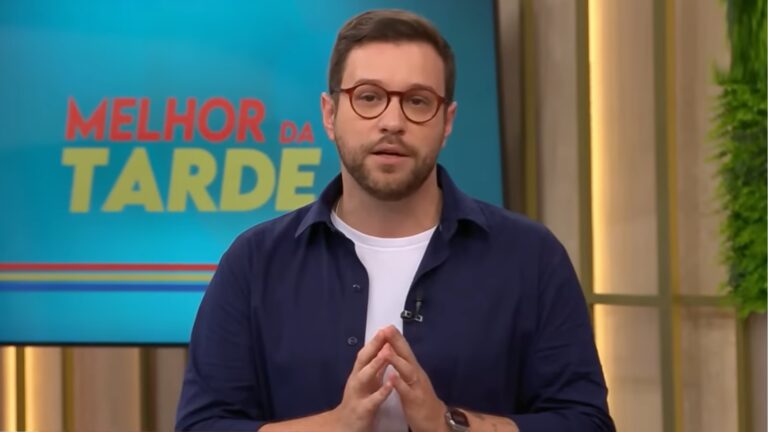 Cassius Zeilmann apresenta o programa Melhor da Tarde, da Band, em estúdio colorido, com o logotipo da atração visível ao fundo. Ele usa óculos de armação redonda, camisa azul sobre camiseta branca e relógio de pulso, falando com as mãos unidas em frente ao corpo